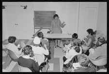 Woman, possibly therapist, standing front people seated chairs during group